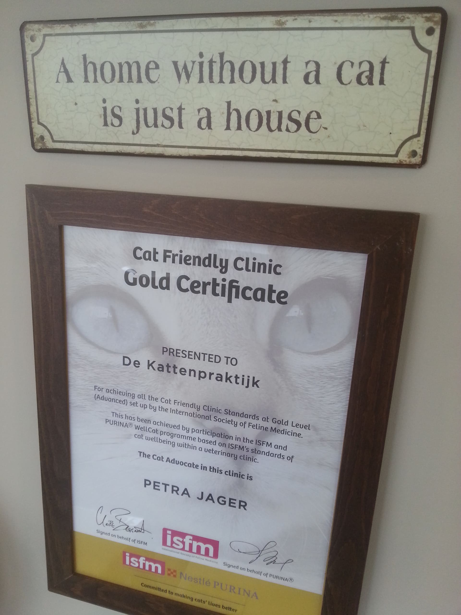 Het ingelijste Cat Friendly Clinic Gold Certificate aan de muur, onder een bordje met de tekst A home without a cat is just a house.