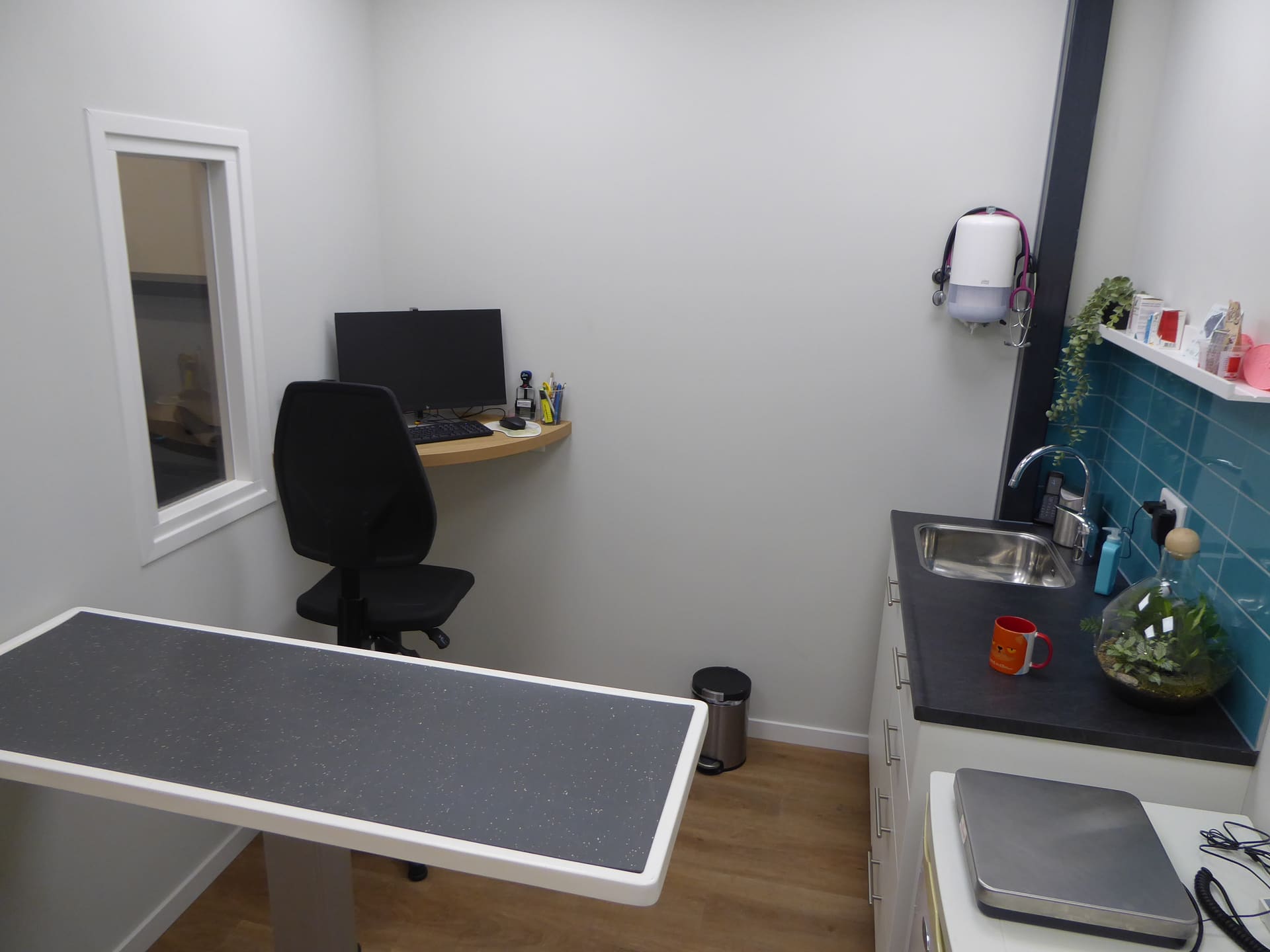 Een spreekkamer met een lage behandeltafel, een doorgeefluik en turquoise tegels boven de spoelbak.